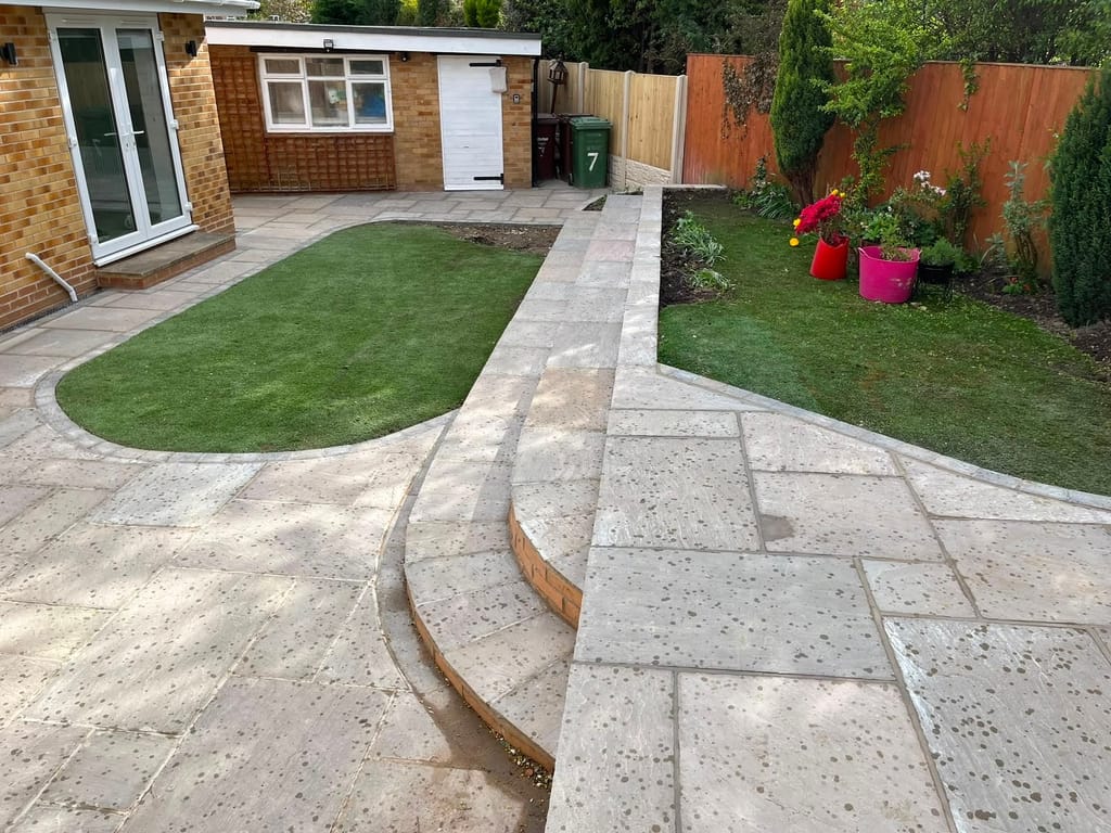 Oval artificial lawn set into cream paving with a brick edged path leading to an outbuilding