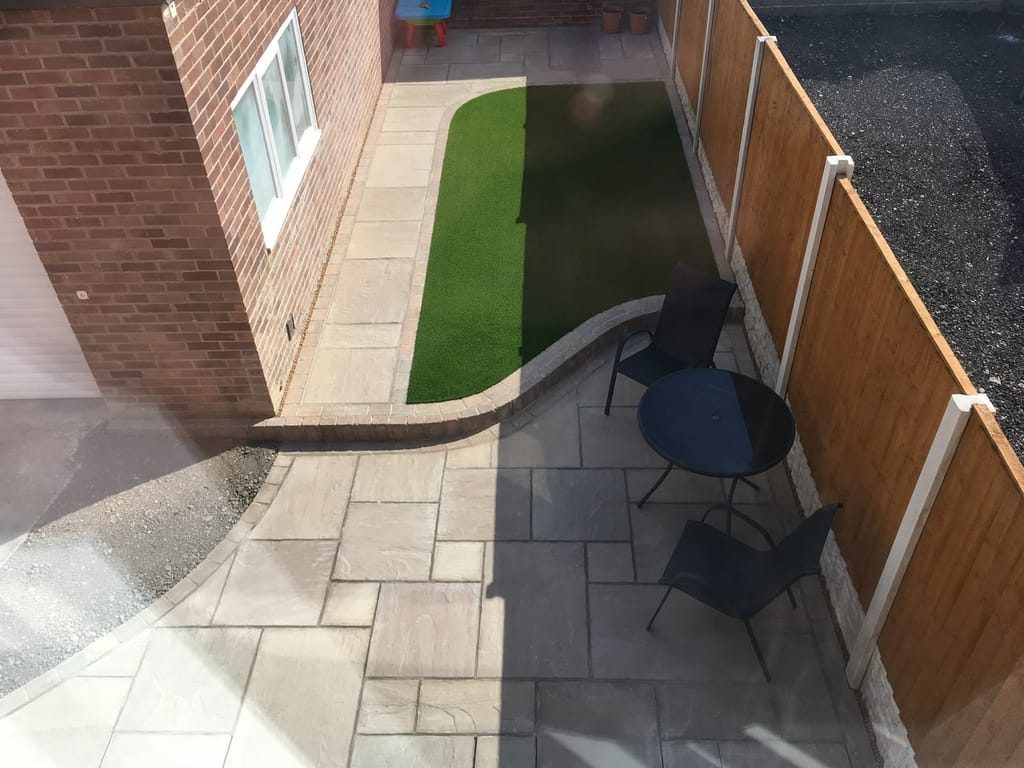 Strip of artificial lawn beside sandstone paving and a low brick wall in a narrow side garden