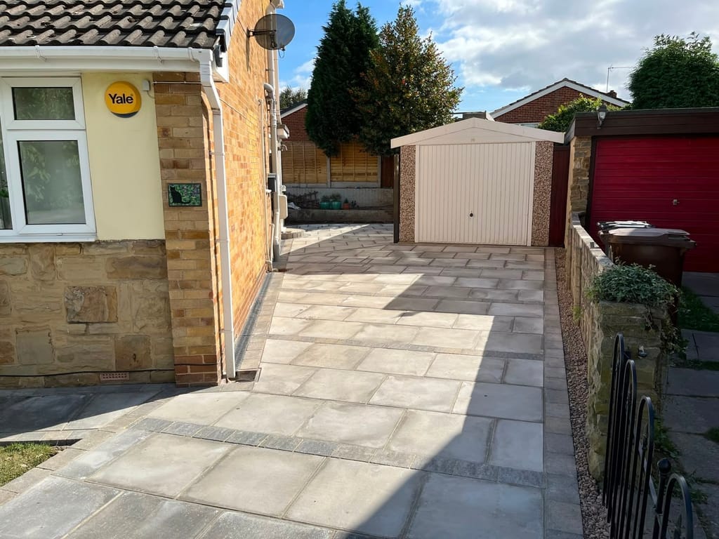 Cream slab paving running from a bungalow frontage to a detached garage past a low stone wall