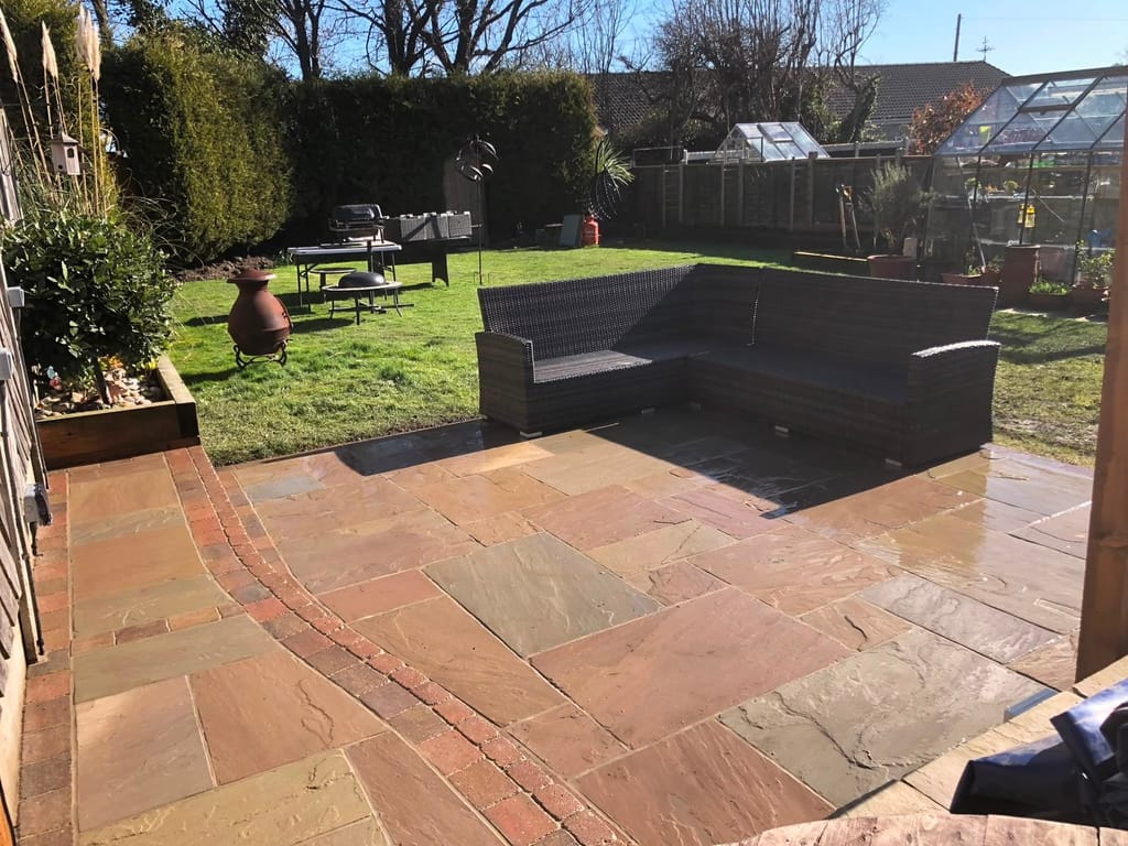 Sandstone patio with a brick banding course and a corner sofa in a long garden with a greenhouse