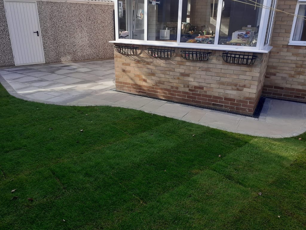 Cream slab patio with a curved edge onto the lawn beside a buff brick bungalow and its garage