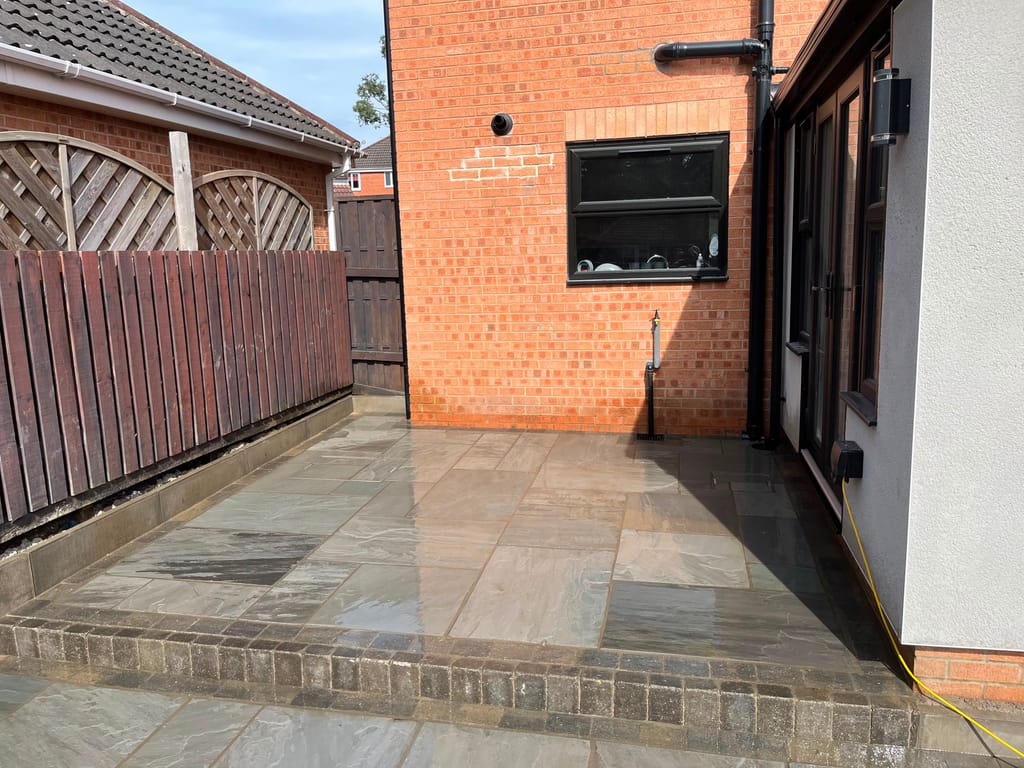 Wet sandstone patio with a sett kerb between a red brick wall and a white rendered extension