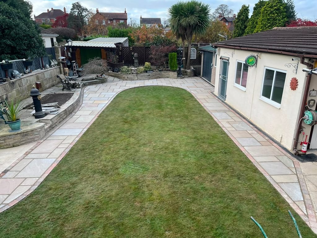 Long garden with sandstone paving down both sides of a lawn past raised beds and an outbuilding