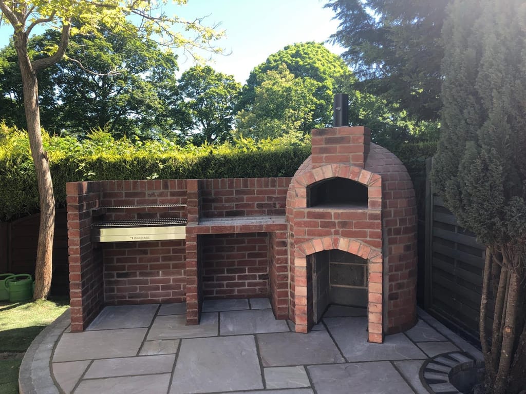 Brick built pizza oven and barbecue counter on grey sandstone paving in a hedged garden